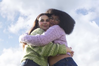 Two friends embrace warmly against a clear blue sky with fluffy clouds, wearing colorful knit
