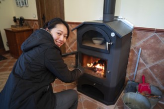 A cheerful Asian woman is focused on managing the fire in a wood burning stove. She is wearing