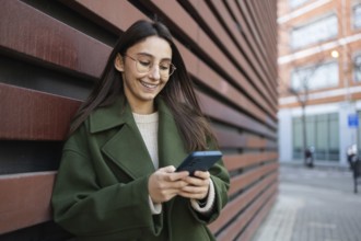 A woman wearing a stylish green coat leans against a wooden wall while engaging with her smartphone