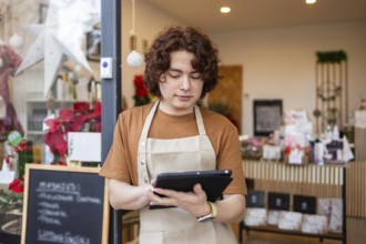 A young male employee stands in front of an organic produce store, wearing an apron and using a