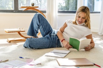 A woman enjoys reading a book while relaxing on a carpeted floor, surrounded by notebooks and a