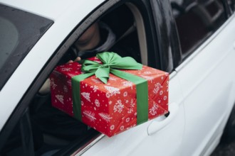 A person hands a festive gift wrapped in red with a green bow from a car window The scene captures