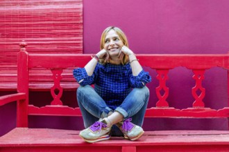 A cheerful young woman in a blue plaid shirt is seated on a bright red bench, smiling and looking