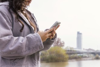 Woman using a smartphone outdoors She appears relaxed, wearing a casual jacket, near an urban river