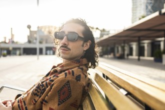 Fashionable man with curly hair and sunglasses lounges on a wooden bench in an urban setting The