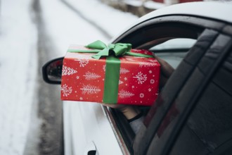 A hand extends a beautifully wrapped gift with red and green holiday paper from a car window The