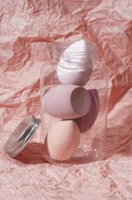 Soft pastel cosmetic sponges in a transparent jar set against a crumpled pink paper background This