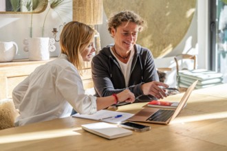 A couple collaborates on a project at home, smiling and engaged They use a laptop and take notes,