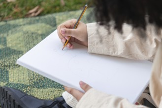 A young black woman sketches on a blank canvas using a pencil, surrounded by grass and natural