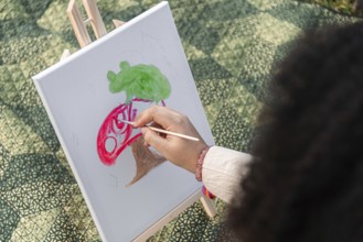 A young black woman skillfully paints a colorful scene on a canvas outdoors. The artwork features