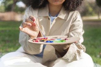 A young black artist holds a paintbrush and a palette filled with vibrant colors, engaging in plein