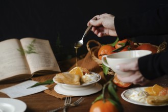 Cropped unrecognizable person drizzling honey over citric crepes, accompanied by fresh orange