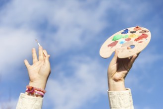 A hand holds a colorful artist palette filled with various paints while another hand reaches up