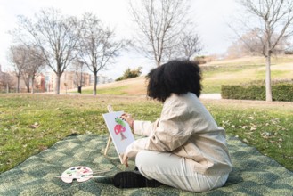A young black woman sits on a blanket in a park, painting a colorful tree on a canvas. Surrounded