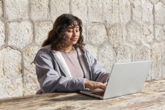 A young Latin woman sitting at an outdoor wooden table, using a laptop Her relaxed expression