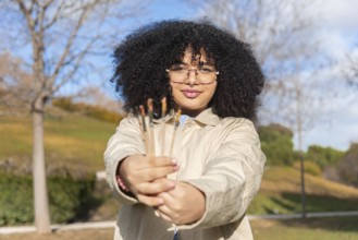 A joyful black woman with glasses and curly hair holds paintbrushes in a sunny park. The background