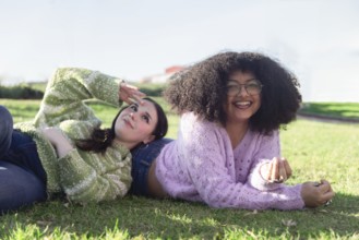 Two friends lie on the grass, enjoying a sunny day in the park. They wear cozy sweaters and smile,