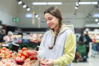 A woman carefully choosing tomatoes in the fresh produce aisle of a busy supermarket. She looks