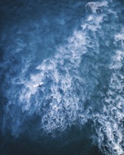 A dramatic aerial shot of a surfer on a white surfboard navigating through foamy blue waves in