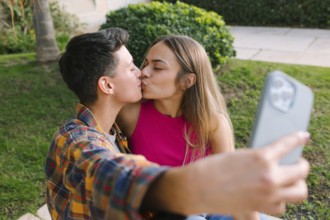 A loving lesbian couple shares a kiss while capturing the moment with a selfie. They sit in a green