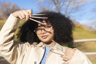 A creative young black artist holding several paintbrushes outdoors, surrounded by nature. The