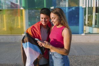 A happy lesbian couple looks at a smartphone together, smiling. One partner is wrapped in a Pride