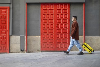 A latino transgender man walks confidently past vibrant red doors, pulling a bright yellow suitcase