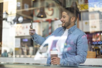 A Latino transgender man in a casual denim shirt smiles while holding a coffee and smartphone at a