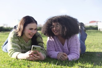 Two friends lie on grass, smiling and enjoying their time while looking at a smartphone in a sunny
