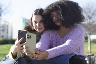 Two friends sitting on grass, taking selfies with their smartphones. They are enjoying a sunny day