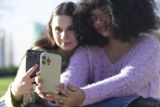 Two friends are sitting outdoors, capturing a selfie with smartphones. They are enjoying a sunny