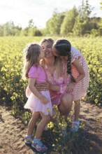 A joyful mother smiles as her daughters kiss her on both cheeks, sharing a tender moment in a