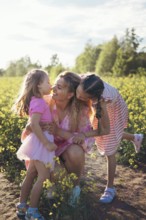 A loving mother kneels in a sunlit flower field as her daughters kiss her cheeks. This heartwarming