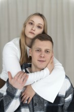 A young man sits as a woman embraces him from behind, both looking calm and affectionate at their