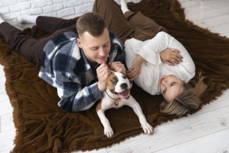 A man and a woman enjoy a playful moment with their dog, reclining on a plush brown blanket in a