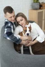 A young man and woman sit closely on a couch, affectionately holding a white and brown