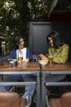 A lesbian couple enjoys drinks at an outdoor cafe. They smile and chat, surrounded by greenery,