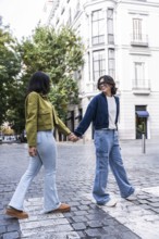 A lesbian couple holding hands while walking on a cobblestone street in the city, surrounded by