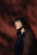 A Latin woman leans against a red wall with her eyes closed, basking in the sunlight. Shadows from