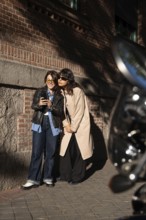 A joyful multiethnic lesbian couple standing in a sunlit urban setting, looking at a smartphone.