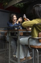 A lesbian couple joyfully toasts with glasses at a cozy cafe patio. They share a moment of laughter