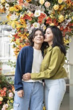 A lesbian couple shares a tender moment in front of a vibrant floral backdrop. Their closeness and