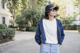 A latin woman wearing a navy cap, sunglasses, white shirt, blue hoodie, and jeans stands with hands