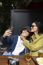 A joyful lesbian couple takes a selfie at a cozy outdoor cafe. They are smiling, surrounded by