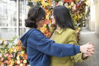 A lesbian couple shares an affectionate moment, embracing amidst a colorful flowered wall. Their