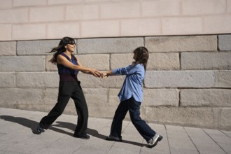A joyful multiethnic lesbian couple holds hands and dances outside against a stone wall,