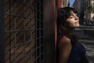 A Latin woman leans against a textured wall with her eyes closed, enjoying the warmth of the sun.