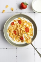 Top down view of a bowl with cornflakes and strawberry slices in milk, with a spoon resting inside.