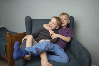Two joyful young brothers share a warm embrace on a cozy blue armchair, radiating happiness and