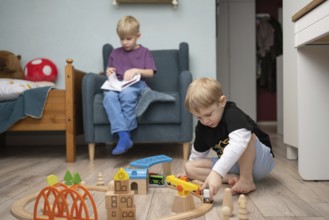 Two young children in a cozy room, one plays with wooden train toys on the floor, while the other